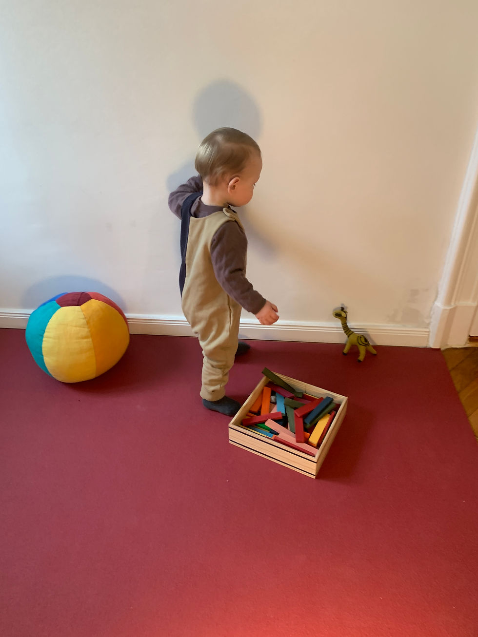 ein junge steht in einem jumpsuit neben einem bunten ball und einer box mit bunten kappla steinen und einer gefilzten giraffe