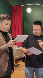 一位西裝主持人與副導演站在室內場景中進行腳本確認，紅色簾幕背景凸顯專業感。