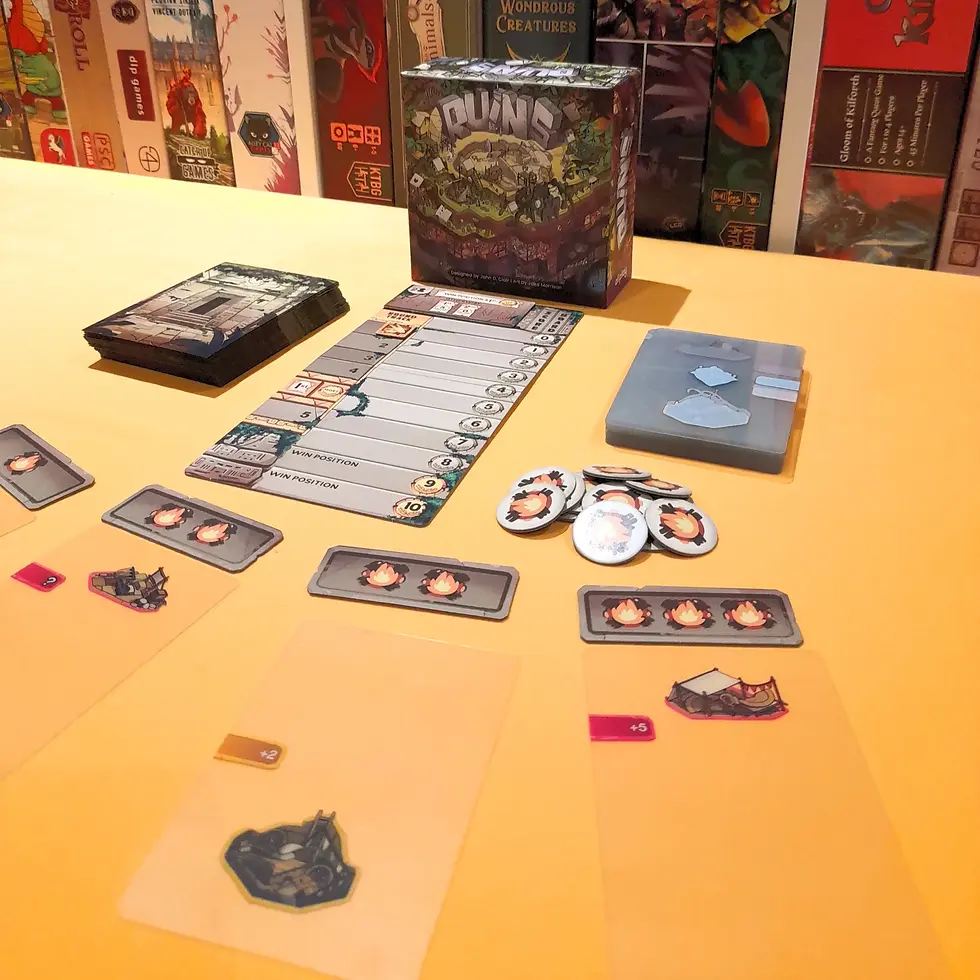Board game setup on yellow table with cards, tokens, and a box titled "Ruins." Background displays a collection of board games.