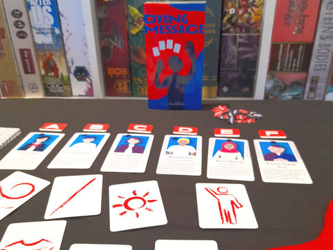 Dying Message board game with components laid out on a black table cloth with board games like Creature Comforts and Wondrous Creatures in the background.