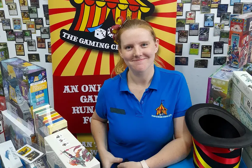 Smiling person in blue shirt at a game store with card backdrop. Colorful boxes and a red-yellow striped hat are on the table.