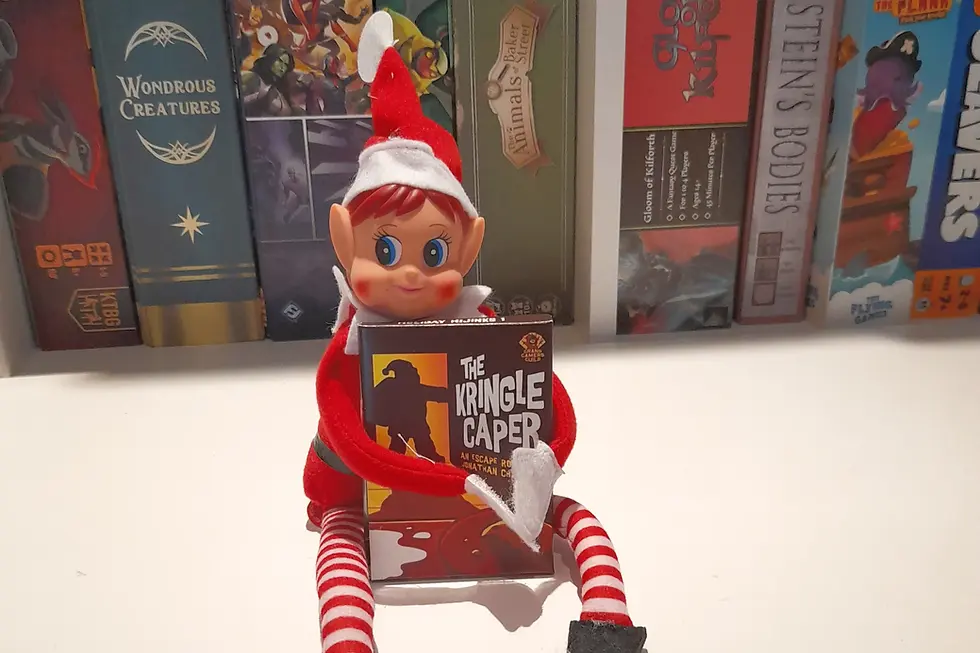 Elf doll in red and white holds "The Kringle Caper" book. Background has board games with colorful spines. Festive and playful scene.
