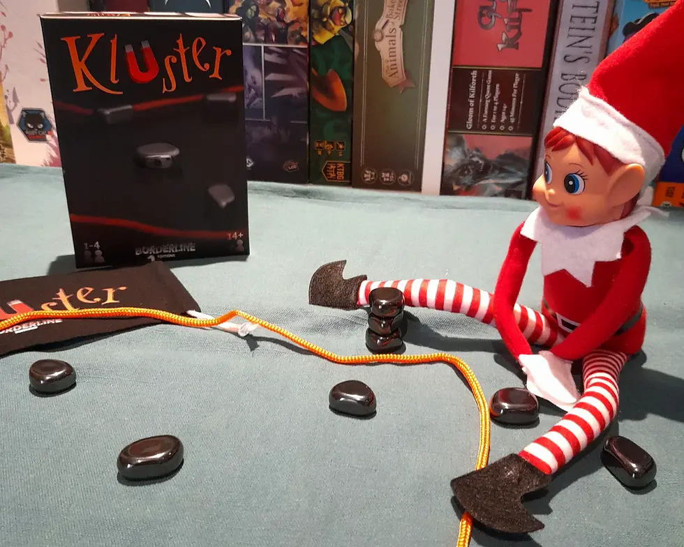 Elf toy in red and white outfit sitting on a teal surface with a magnetic game, "Kluster" box, and orange string around black stones.