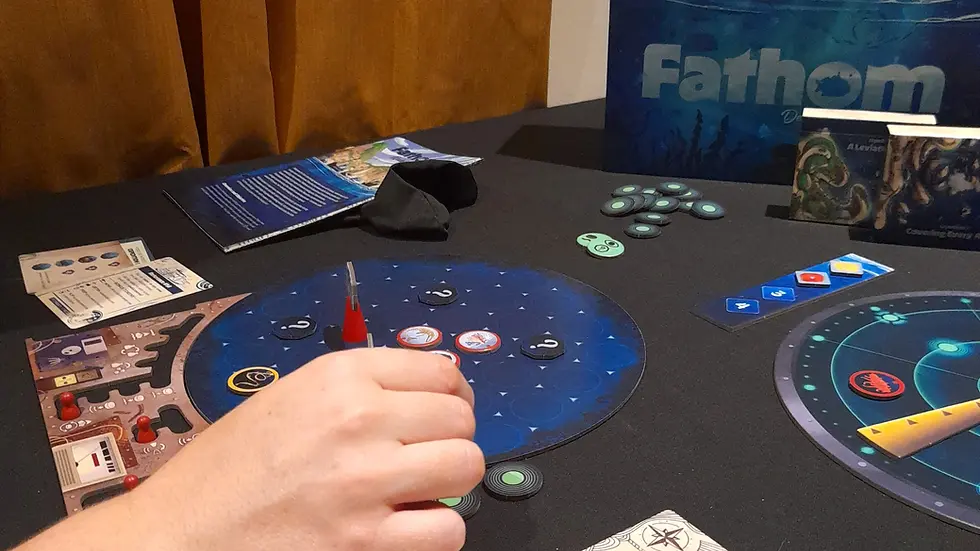 Hand moving tokens on a blue board game titled "Fathom" with various cards and pieces on a dark table. Cards feature sci-fi designs.