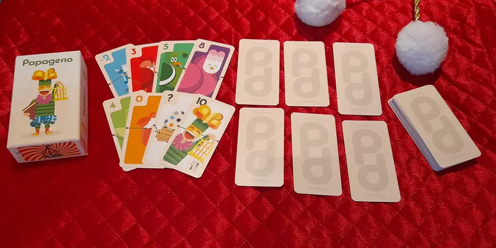 Papageno card game set on red quilted fabric. Colorful cards display various numbers and characters. Box and cards have "Papageno" text.
