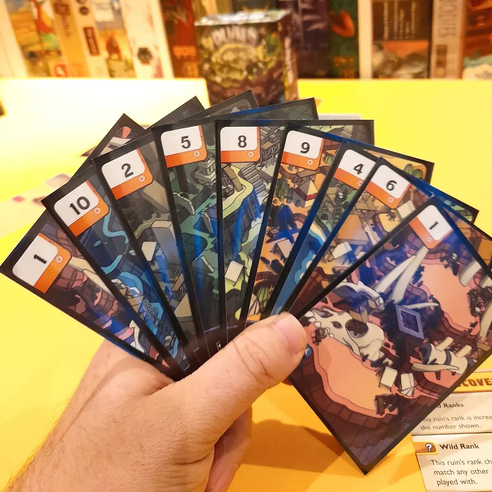 A hand holds colorful numbered cards, each with unique illustrations, against a bright yellow table. Blurred board game boxes are in the background.