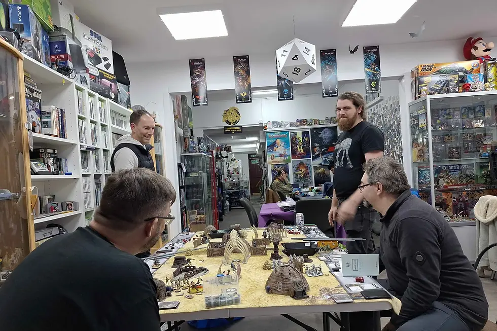 Four men play the Star Wars Legion tabletop game in a well-lit game store. Posters and game boxes fill the shelves. A large dice hangs from the ceiling.