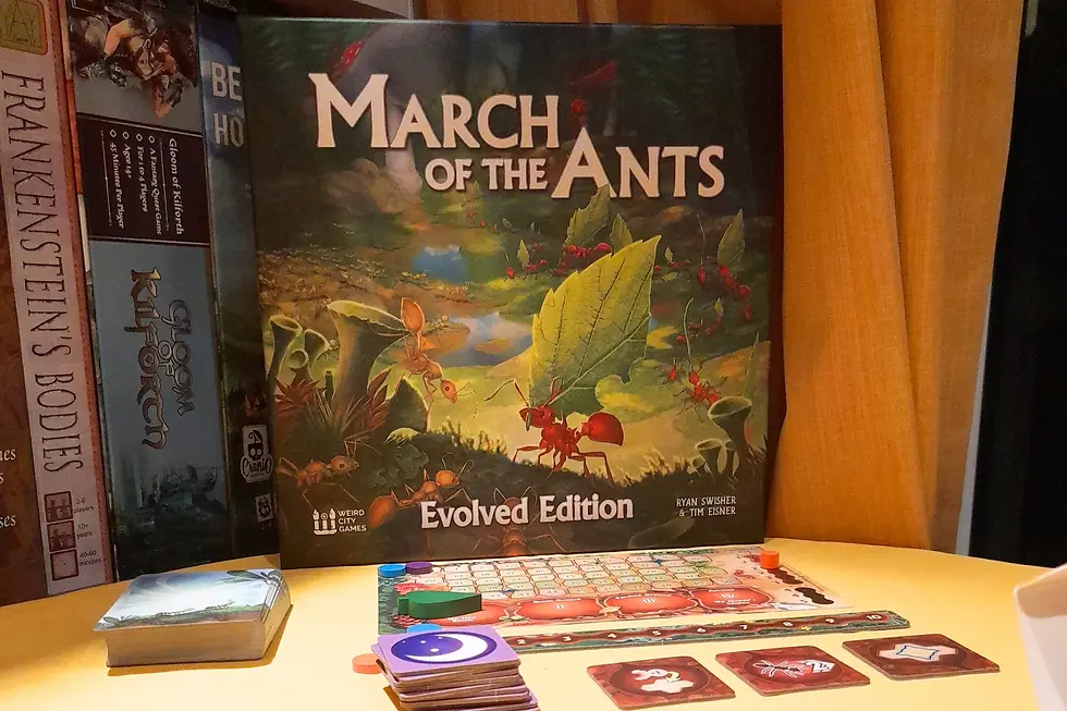 "March of the Ants" Evolved Edition board game cover with ants and leaves. Nearby are cards and game pieces on a yellow surface.