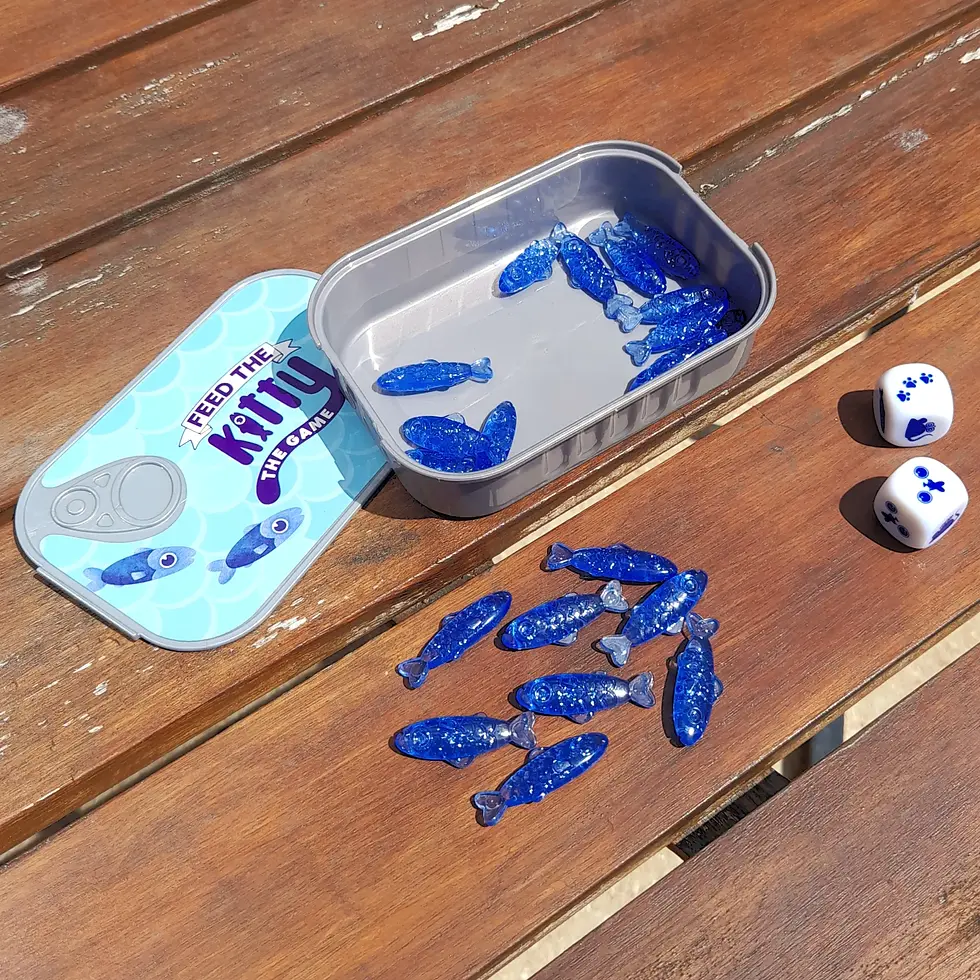 Open tin with blue fish-shaped pieces, dice on a wooden table. Lid reads "Feed the Kitty: The Game." Sunny day, relaxed mood.