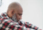 Bald man with a gray beard in a red plaid shirt, looking down pensively against a light background.