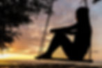 Silhouette of a person sitting on a swing at sunset, with a calm ocean and trees framing the scene. The mood is peaceful and reflective.