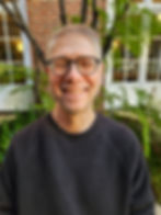 Portrait of David Lesseps in front of windows and a brick background, black top, smiling, plants