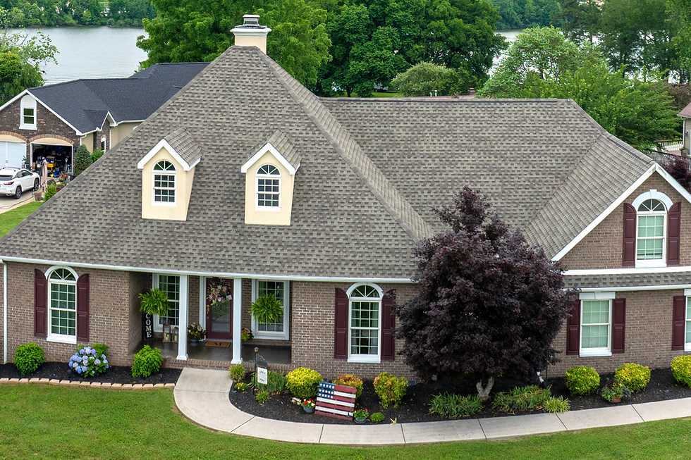 Brick house with a large gray roof, arched windows, and a manicured garden. An American flag pallet decorates the front yard. Trees and a lake are in the background.