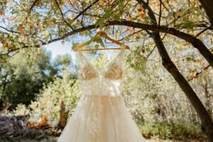Photo of a werdding dress hanging by a hanger on a tree branch. Surrounded by green trees and sunshine. Captured by Denver wedding photographer, Michelle Betz Photography
