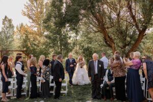 bride walking down isle with father. The guests are standing watching. Surrounded by green grass and trees and sunshine
