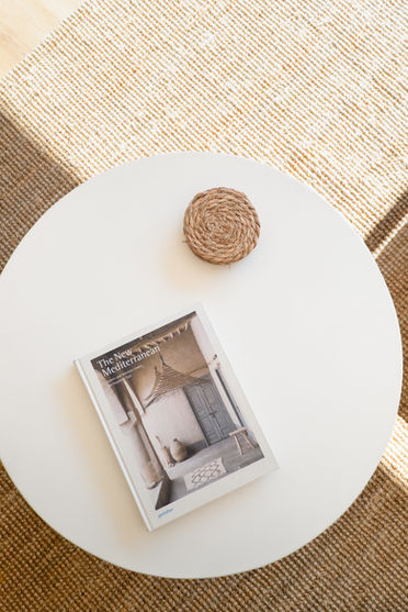 Vista cenital de una mesa auxiliar blanca con un libro de diseño y un posavasos de fibra sobre alfombra de yute.