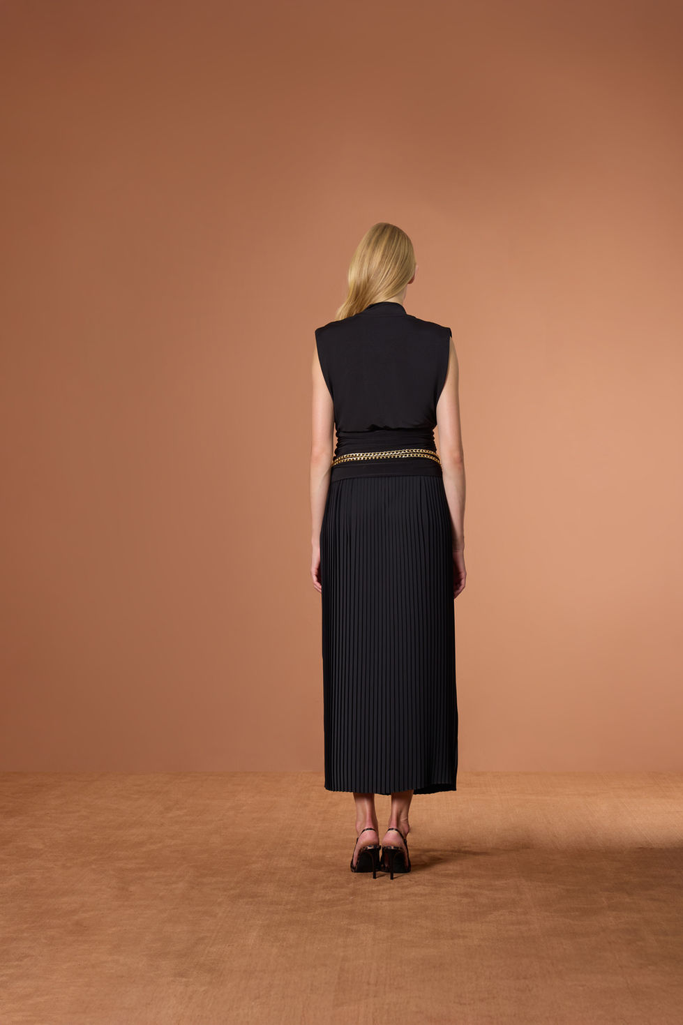 Kadın arkadan siyah kolsuz üst, uzun etek, altın kemerle poz veriyor.