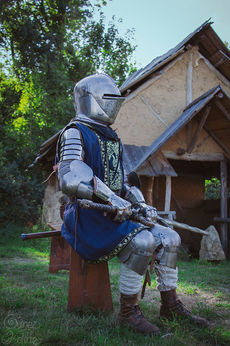 Photographie en format portrait d’une homme portant une armure de la tête aux pieds. Il est assis sur un banc. Il tient une épée longue posée sur ses genoux. Par dessus son armure, il porte un habit bleu qui descend jusqu’au bas de ses cuisses. Il y a des broderies dorées tout le long des bords et au niveau du torse. Derrière lui, il y a une maison fait en torchis avec le toit construit par des planches de bois. Elle sort du cadre à droite. Derrière le bâtiment, à gauche, il y a des arbres. De l’herbe habille le sol de partout.