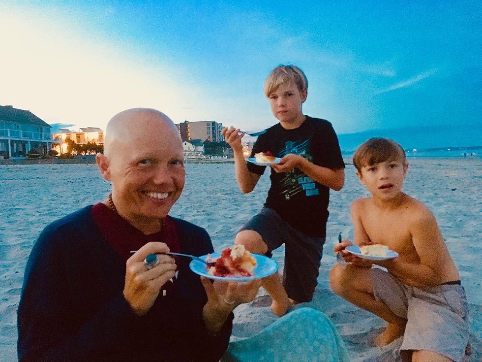 Author with bald head and two sons eat cake on the sand by the ocean at dusk.