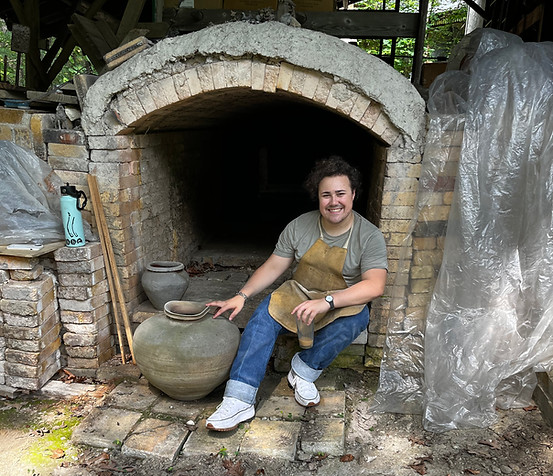 Gabriel Cordero sitting at the mouth of Joy browns anagama kiln, smiling into the camera wile holding a cup of iced coffee in one hand, and resting his other hand on a large pot 