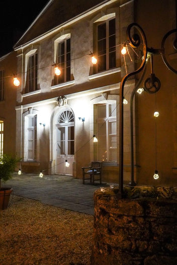 Evening view of Château Chevreuil's stone exterior with warm lighting, hanging festoon bulbs, and a rustic stone well in the courtyard.
