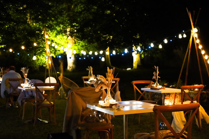 Enchanting outdoor night event at Château Chevreuil with lit-up tables, wooden chairs, and hanging string lights in a garden setting.