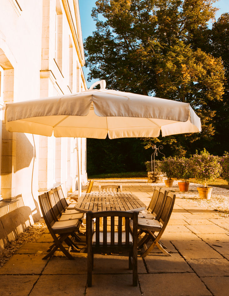 A wooden table with chairs set on the stone patio of the castle, shaded by a large umbrella.