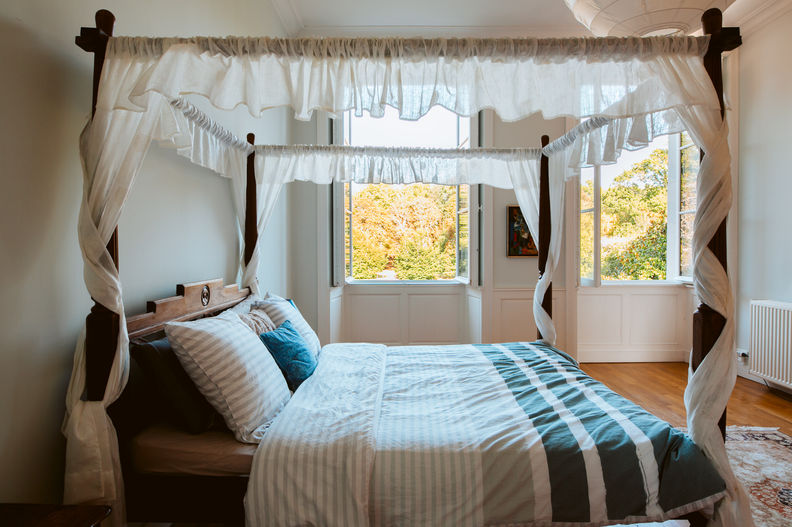 The queen’s room four-poster bed with its white fabric curtains in a room with a window. The bed has a beautifully made wooden frame.
