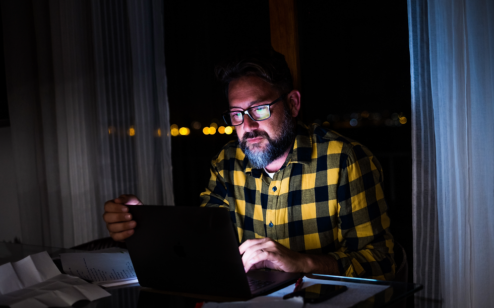 man sitting in dark room at night holding and working on laptop at desk with paper about