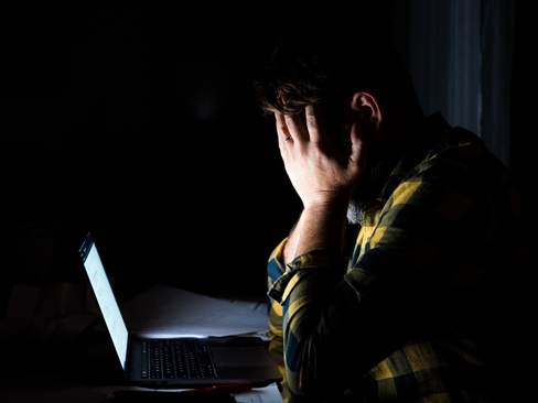 man sitting at his desk in the dark at night with hands pressed up to face covering self stressed