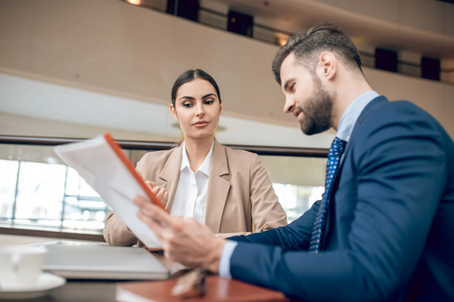 Two professionals sit at a table, reviewing documents. The woman listens intently. They're in a modern office with neutral tones.