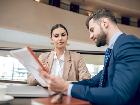 Two professionals sit at a table, reviewing documents. The woman listens intently. They're in a modern office with neutral tones.