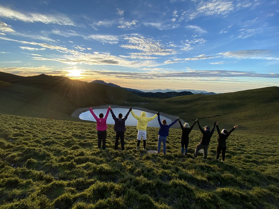 7 people joining hands in the air at the top of a mountain at sunrise facing a lake in front of them