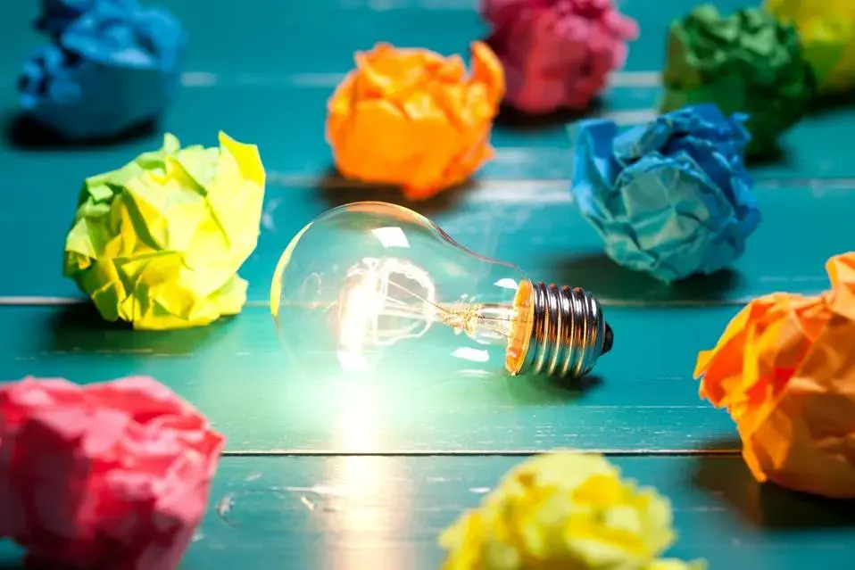 A light bulb on a table with balled up papers of various colors.