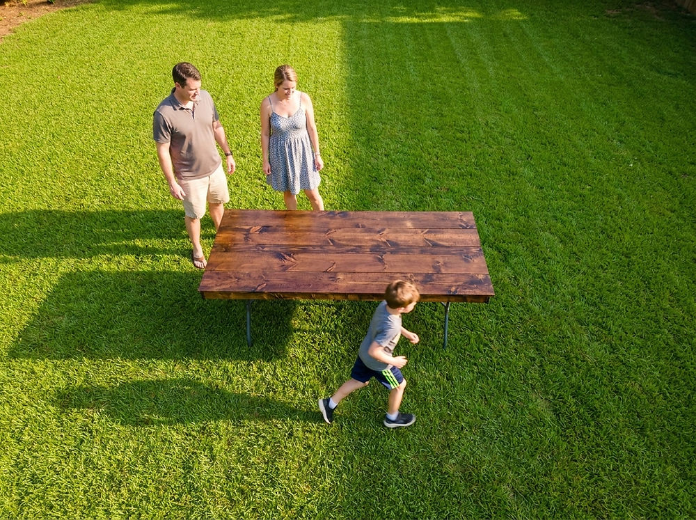 A low aerial shot of a backyard with green grass and a family standing near a wood top table. 