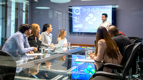 Image/Canva Alt-txt: A group of people in a conference room watching a presentation on a large screen