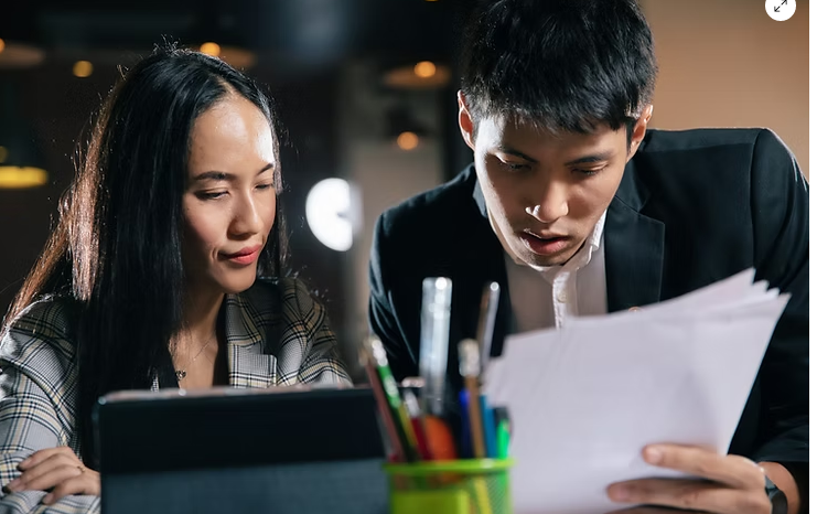 A man and woman reviewing documents together at a table with a green pen holder and a device.