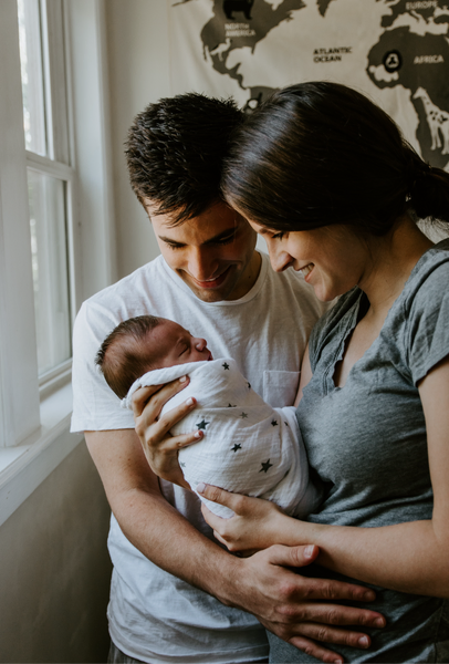 Smiling parents holding their newborn baby by a window – representing early bonding and sleep support during the fourth trimester.