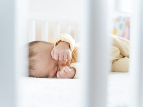 Baby sleeping in a crib, wearing a striped onesie, arms raised near face. Soft lighting and white bedding create a peaceful mood.