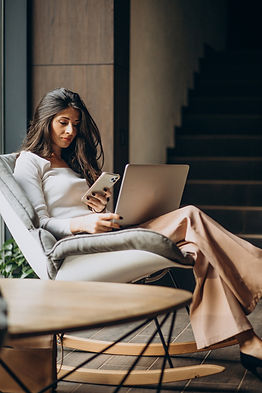 Young woman sitting on a chair with laptop on her lap and reading from phone