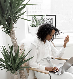 Woman in a white outfit working on a laptop at home, seated beside a tall indoor plant and large window, creating a calm and focused workspace.