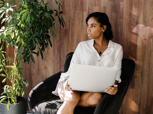 Woman in white shirt sitting on black chair with a laptop, next to a green potted plant. Wooden wall background, natural light.
