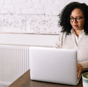 A woman with curly hair and glasses types on a laptop at a wooden table. White textured wall in the background, cup nearby.
