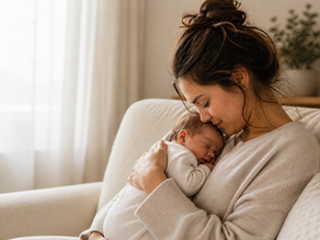 A woman gently cradles a sleeping baby, their foreheads touching, on a beige sofa with soft lighting and a plant in the background.