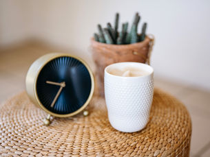 A white mug, a black and gold clock, and a cactus in a pot sit on a woven surface. Neutral tones create a calm, cozy mood.
