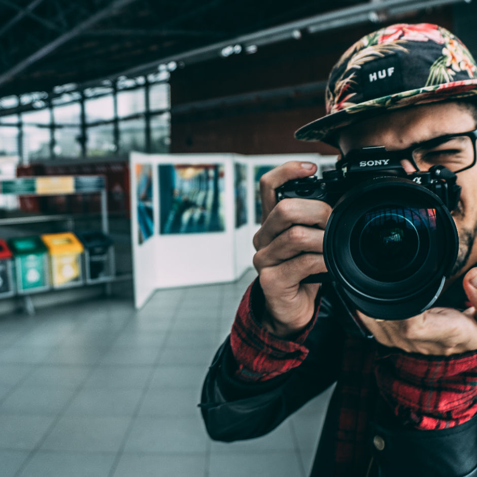 A person wearing a floral cap and red checkered shirt holds a Sony camera in an indoor setting with colorful recycling bins.