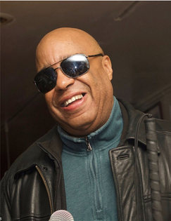Frank smiling between verses, holding a microphone. Wearing shades, a dark green turtleneck and leather jacket.