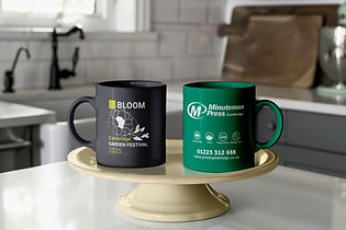Two coloured printed mug, one grey and one green, displayed on a cake stand on kitchen counter top. Green mug printed with Minuteman Press Cambridge logo in white along with business icons, contact number and website address. Grey mug printed with Cambridge Garden Festival design in white and yellow.