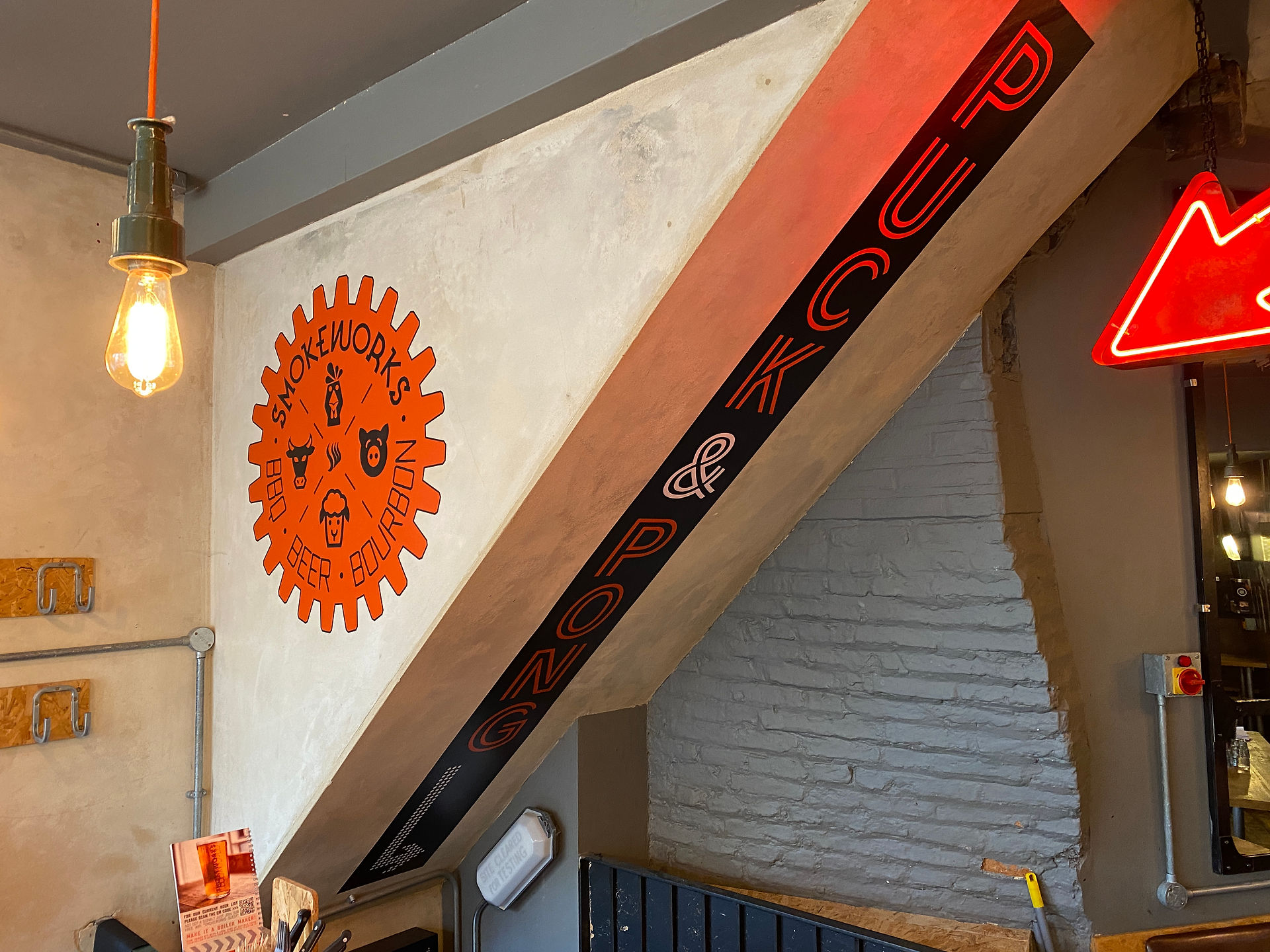 Printed Vinyl Wall Graphics shown installed shown installed in Cambridge Smokeworks restaurant. Orange icon logo graphic in cogwheel shape displayed on far wheel, vertically typed 'Puck & Pong' displayed on ceiling above stairs leading to the downstairs of restaurant. Wall painted with a distressed yellow and beige effect.