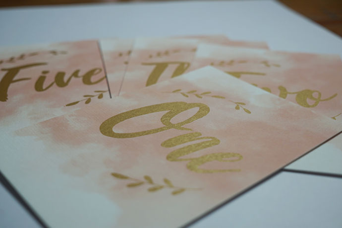 Printed table numbers using a hand written effect font with Metallic Gold on pink watercolour background.
